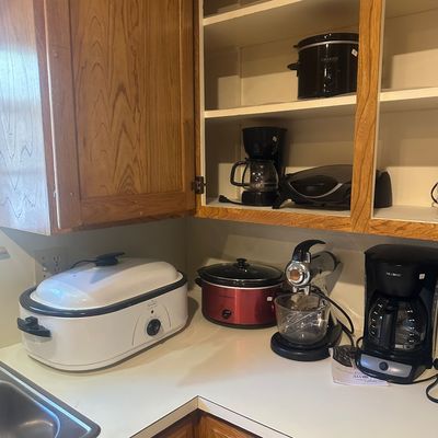 Sale Photo Thumbnail #38: Assortment of small kitchen appliances, including coffee makers, slow cookers, a stand mixer, and a Rival roaster oven arranged on a countertop and in oak cabinets.