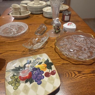 Sale Photo Thumbnail #36: Collection of tabletop glassware, ceramics, and serveware arranged on a wooden table, including clear pressed glass platters, a hand-painted fruit square plate, china dinnerware stacks, and decorative figurines.
