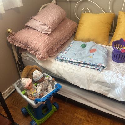 Sale Photo Thumbnail #65: White metal daybed styled with pink ruffled pillows, gold cushions, and a folded baby blanket, accompanied by a plastic toy shopping cart with dolls.