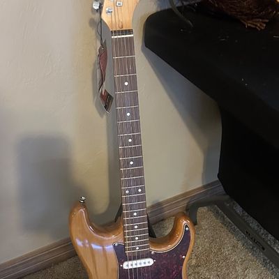 Sale Photo Thumbnail #33: Natural wood electric guitar with tortoise shell pickguard, three single-coil pickups, and chrome hardware, resting on a floor stand.