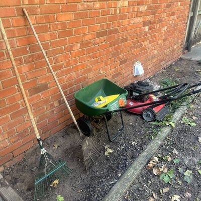 Sale Photo Thumbnail #23: Collection of lawn care equipment including two metal leaf rakes, a green lawn spreader, and a red walk-behind lawn mower resting against a brick exterior wall.
