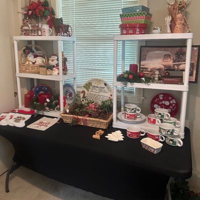 Sale Photo Thumbnail #6: Collection of Christmas decor and tableware displayed on a black-covered table and white plastic shelving units, featuring mugs, decorative plates, figurines, and holiday tins.