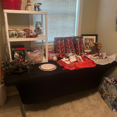 Sale Photo Thumbnail #52: Collection of Christmas decorations including boxed red glass ornaments, holiday mugs, figures, and craft items displayed on a black tablecloth and plastic shelving unit.