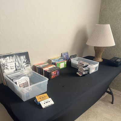 Sale Photo Thumbnail #56: Collection of media including vinyl record albums, cassette tapes, CDs, and VHS tapes arranged on a table with a pleated lamp.