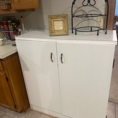 Sale Photo Thumbnail #42: White two-door storage cabinet featuring silver handles, topped with a framed art piece and a black wrought iron scrollwork rack.