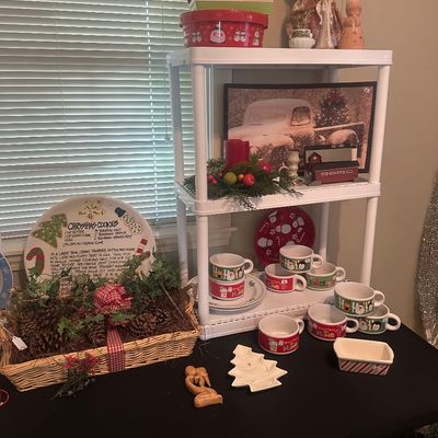 Sale Photo Thumbnail #43: Collection of holiday table decor and servingware, including a pinecone basket display, festive ceramic mugs, decorative tins, and angel figurines displayed on a white shelving unit.