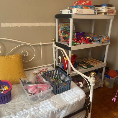Sale Photo Thumbnail #66: White metal frame daybed with brass finials alongside a plastic shelving unit filled with board games, toys, and puzzles.