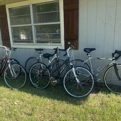 Sale Photo Thumbnail #2: Collection of adult bicycles lined up against a house, including a silver Cannondale road bike and hybrid models from Giant and Schwinn.