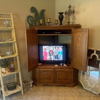 Sale Photo Thumbnail #26: Traditional oak corner media armoire with lower cabinet storage, housing a flat-screen television and flanked by a distressed ladder shelf unit and an accent mirror.
