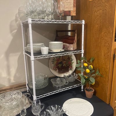 Sale Photo Thumbnail #39: A collection of glassware, ceramic dishware, and decorative items displayed on a white wire shelving unit and table, including wine glasses, crystal bowls, and a faux potted lemon plant.