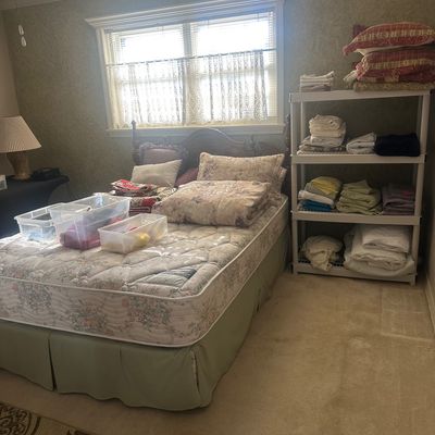 Sale Photo Thumbnail #54: Bedroom featuring a double bed with a wooden headboard, floral mattress, and plastic storage tubs, alongside a plastic shelving unit stocked with folded linens and pillows.