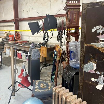 Sale Photo Thumbnail #52: Eclectic collection of items in a warehouse setting, including studio stage lights on stands, an ornate grandfather clock, audio equipment, and a Chinese lacquered folding screen.