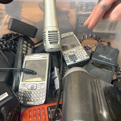 Sale Photo Thumbnail #21: Clear plastic storage bin filled with vintage electronics, including a silver handheld microphone, Palm Treo smartphones, Motorola flip phones, and chargers from the early 2000s.