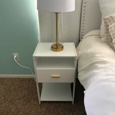 Modern white nightstand featuring a fluted drawer front, open shelf, and gold-tone hardware, topped with a contemporary clear glass and...