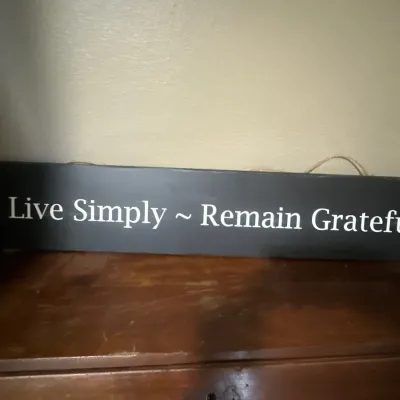 Black wooden wall plaque reading "Live Simply ~ Remain Grateful" in white lettering