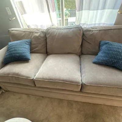 Contemporary three-cushion sofa upholstered in a light taupe microfiber fabric, featuring rounded armrests and accompanied by two blue...