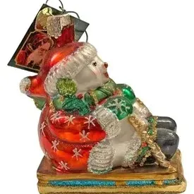 Blown glass Christmas ornament featuring a snowman riding a sled, detailed with glitter accents and original tag attached.