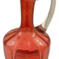 Vintage red glass pitcher featuring a panel-cut bulbous body, narrow neck, ruffled spout, and a clear applied glass handle.