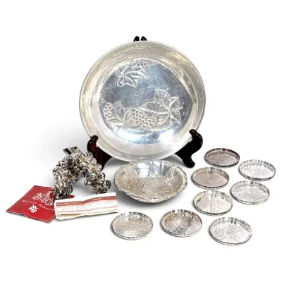 Sale Photo Thumbnail #177: Collection of silver-plated tableware featuring an embossed grape-and-vine platter on a wooden stand, a small floral dish, a set of engraved glass coasters with metal rims, and silver-plated napkin rings with WMF paperwork.