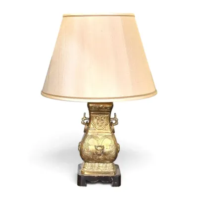 Sale Photo Thumbnail #185: Vintage Asian-style cast brass table lamp featuring ornate relief motifs and loose ring handles, mounted on a dark wood base with a beige fabric shade.