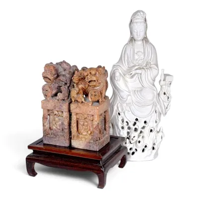 Sale Photo Thumbnail #233: A pair of intricately carved soapstone foo dog bookends on a wooden stand alongside a Blanc de Chine porcelain figure of Guanyin.