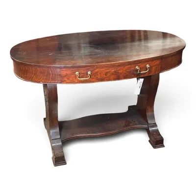 Sale Photo Thumbnail #129: Antique quarter-sawn oak library table featuring an oval top, single wide drawer with brass bail handles, and a curved stretcher base.