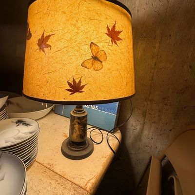 Sale Photo Thumbnail #116: Small Oriental-style table lamp featuring a dark base with gilt landscape details, paired with a illuminated paper drum shade embellished with pressed leaves and a butterfly motif.