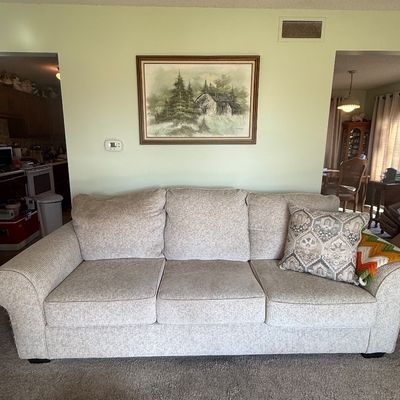 Sale Photo Thumbnail #60: Contemporary three-cushion upholstered sofa in light beige heather fabric, accompanied by patterned accent pillows and a framed landscape print overhead.