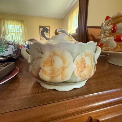 Sale Photo Thumbnail #43: Hand-painted porcelain covered tureen featuring large yellow roses and gold-accented handles, resting on a wooden tabletop.