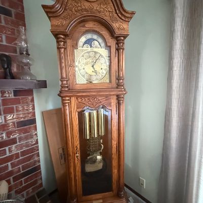 Sale Photo Thumbnail #91: Traditional oak grandfather clock featuring carved floral bonnet top, brass pendulum, chime weights, and a moon phase dial.