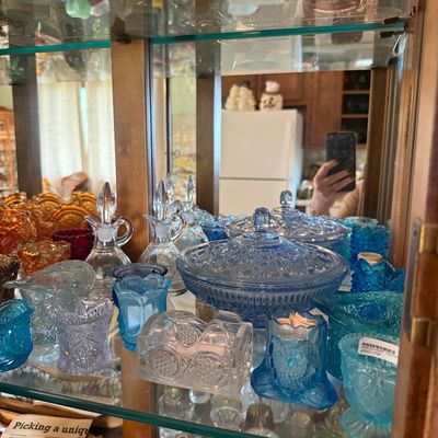 Sale Photo Thumbnail #94: Collection of vintage colored pressed glass and glassware display items, including blue and amber dishes, decanters, and toothpick holders inside a mirrored display cabinet.