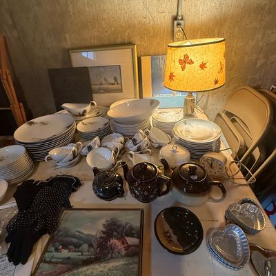 Sale Photo Thumbnail #115: Assortment of vintage tabletop items including a large dishware set, ceramic teapots, a framed landscape painting, heart-shaped tin baking molds, and a table lamp with a botanical shade.