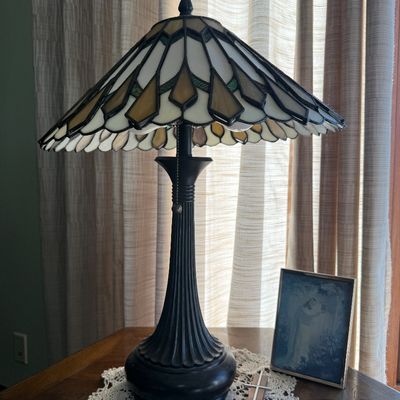 Sale Photo Thumbnail #61: Tiffany-style table lamp with a geometric leaded slag glass shade in cream, amber, and green tones, set on a dark metal ribbed base.