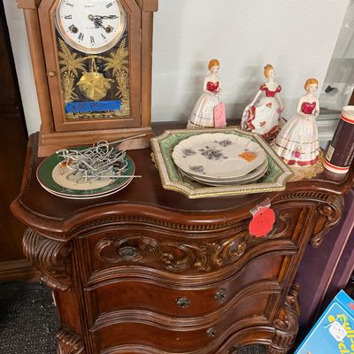 Traditional carved wood bombé nightstand display featuring a Formac mantel clock, porcelain lady figurines, and decorative plates.