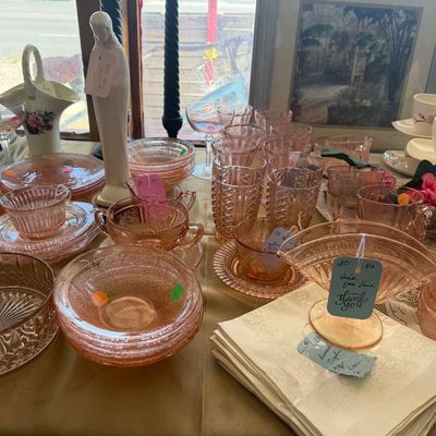 Collection of vintage pink depression glass glassware, including a fan vase, teacups, saucers, and tumblers displayed alongside a white...