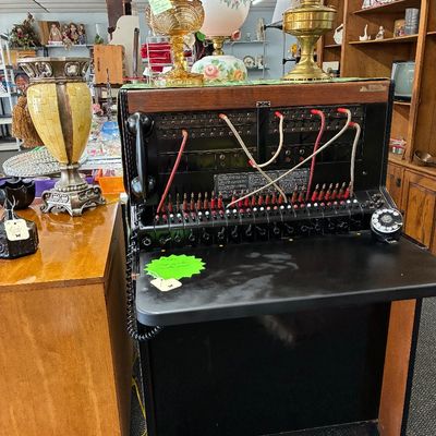 Vintage wood-cased telephone switchboard console with rotary dial and patch cords, displayed alongside various antique lamps and home decor.