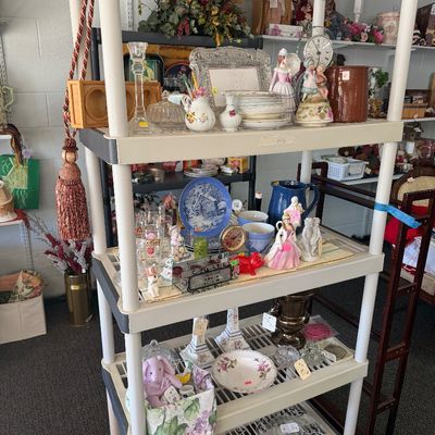 White plastic four-tier storage rack filled with various vintage home decor, porcelain figurines, glassware, and ceramic dishware.