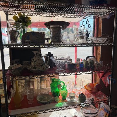 Wire shelving unit filled with vintage glassware, ceramic teacups and pitchers, decorative bowls, and green depression glass.