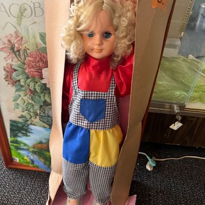 Vintage blonde doll with curly hair, wearing a red shirt and colorful primary-patterned overalls, housed in its original damaged box.