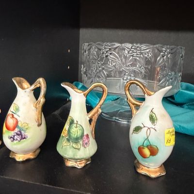 Trio of vintage miniature porcelain pitchers featuring hand-painted fruit motifs and gilded accents, displayed in front of a clear glass...