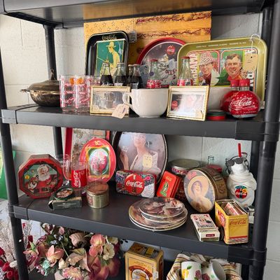 Black plastic shelving unit filled with vintage Coca-Cola memorabilia, including serving trays, glass bottles, collectible tins, drinking...
