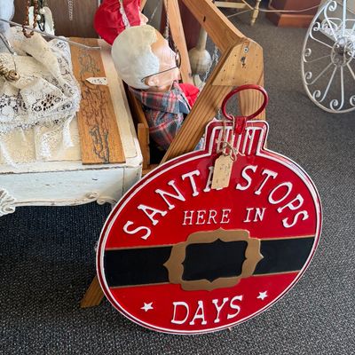 Red metal round 'Santa Stops Here In... Days' countdown sign leaning against an A-frame wooden display with vintage dolls...