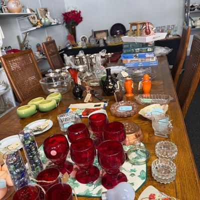 A large dining table displayed with a variety of vintage glassware, ruby red goblets, silver-plated items, ceramics, and home decor.