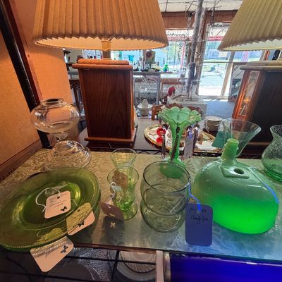 Collection of vintage green glassware, uranium glass pieces, and clear oil lamps displayed on a shelf beneath a pair of wooden table lamps.