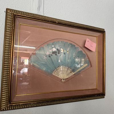 Framed vintage hand fan featuring painted floral details on light blue feathers with a carved bone or faux-bone structure, presented in a...