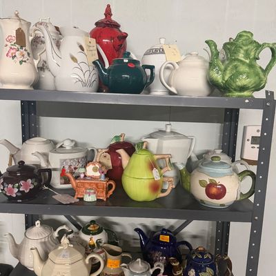 Collection of decorative ceramic teapots displayed on a three-tier metal shelf unit, featuring various novelty, floral, and fruit-shaped...