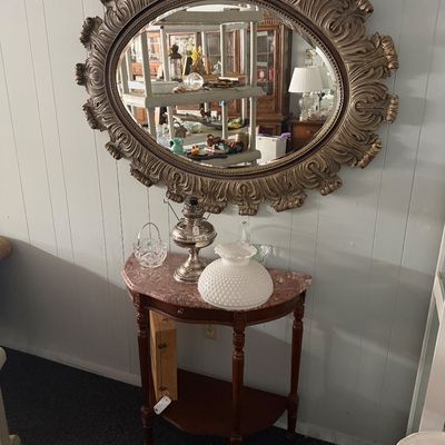Large ornate gold-toned oval mirror wall-mounted above a marble-top half-moon console table, displaying vintage glass decor and a...