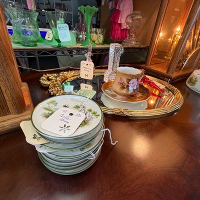 Collection of vintage decorative glassware and porcelain, featuring a stack of small scenic coasters, a floral teacup and saucer, and...