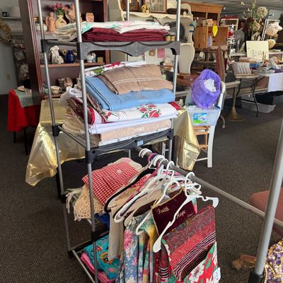 Metal and plastic multi-tier garment rack filled with folded household linens, tablecloths, and hung decorative fabrics.