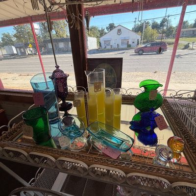 Collection of vintage colored glassware, including pitchers, vases, tumblers, and dishes, displayed on a wrought-iron cart with a...